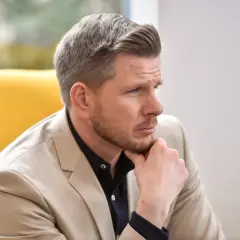 Mężczyzna w beżowej marynarce i granatowej koszuli, z ręką na brodzie, zastanawia się nad pytaniem: ile lat ma Filip Chajzer?