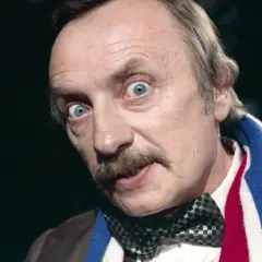 Mężczyzna z niebieskimi oczami i wąsami, w muszce i szaliku w barwach narodowych. Wygląda, jakby ogłaszał: "poszukiwany poszukiwana obsada!".