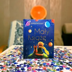 Odkryj, dlaczego warto przeczytać Małego Księcia. Książka z liskiem i chłopcem na tle kosmosu, otoczona rozsypanymi puzzlami.