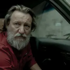 Wzgórze psów obsada: Ian McShane w czerwonej koszuli siedzi w samochodzie, z siwą brodą i zmęczonym spojrzeniem.