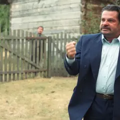 Mężczyzna w garniturze, jakby z filmu "To ja, złodziej", pokazuje pięść. Za nim przez płot wyglądają inni.