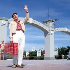 Truman Show obsada: Jim Carrey wita się z widzami, trzymając czerwoną walizkę. W tle łuk z napisem "Omnibus".