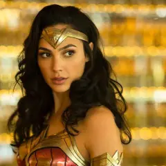 Gal Gadot jako Wonder Woman, część obsady filmu, w złotej zbroi i opasce na głowie, na tle rozmytych, złotych świateł.