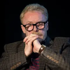 Daniel Olbrychski w okularach, z dłońmi złożonymi na brodzie. Choć nie widać tego na zdjęciu, ile wzrostu ma Daniel Olbrychski, jego postawa sugeruje pewność siebie.
