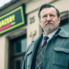 Mężczyzna w eleganckim swetrze i płaszczu, z siwą brodą, stoi przed budynkiem z neonem. To jeden z aktorów z obsady "Minuta ciszy".