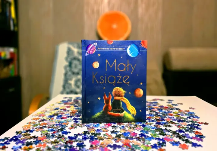 Odkryj, dlaczego warto przeczytać Małego Księcia. Książka z liskiem i chłopcem na tle kosmosu, otoczona rozsypanymi puzzlami.