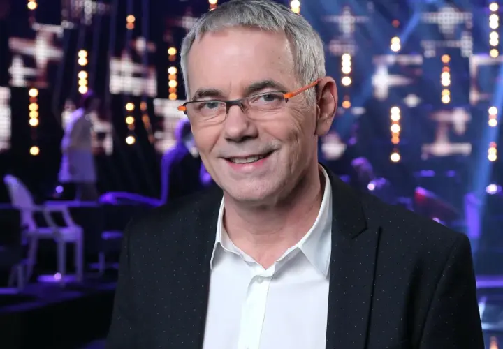 Uśmiechnięty Robert Janowski w okularach na tle sceny. Wiele osób zastanawia się, ile lat ma Robert Janowski, ale jego wiek nie jest widoczny na zdjęciu.