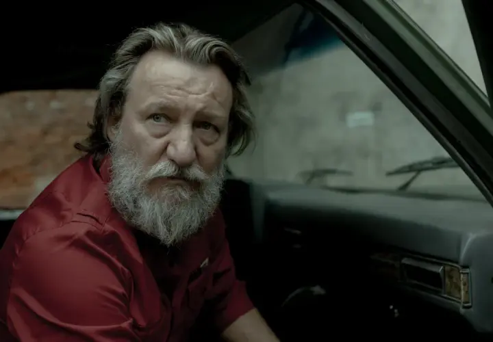 Wzgórze psów obsada: Ian McShane w czerwonej koszuli siedzi w samochodzie, z siwą brodą i zmęczonym spojrzeniem.