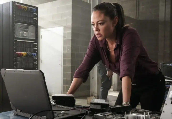 NCIS Hawaii obsada: Jane Tennant analizuje dane na laptopie, otoczona serwerami i dyskami twardymi.