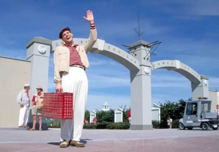 Truman Show obsada: Jim Carrey wita się z widzami, trzymając czerwoną walizkę. W tle łuk z napisem "Omnibus".