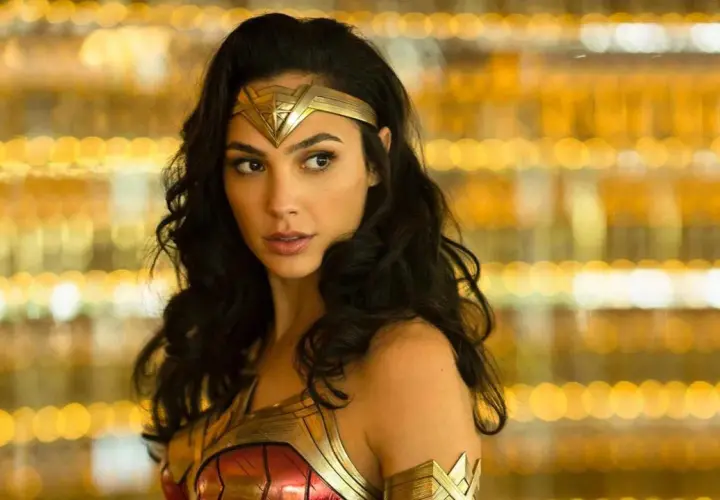 Gal Gadot jako Wonder Woman, część obsady filmu, w złotej zbroi i opasce na głowie, na tle rozmytych, złotych świateł.