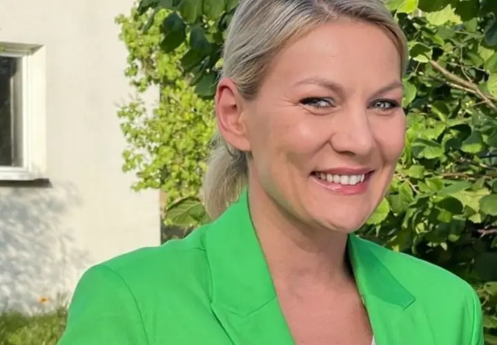Uśmiechnięta Martyna Kupczyk w zielonej marynarce, na tle zieleni i białego domu.