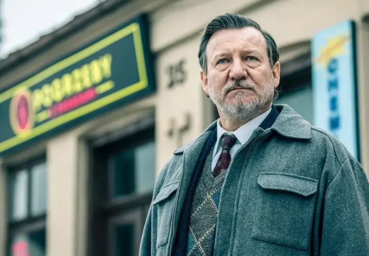 Mężczyzna w eleganckim swetrze i płaszczu, z siwą brodą, stoi przed budynkiem z neonem. To jeden z aktorów z obsady "Minuta ciszy".