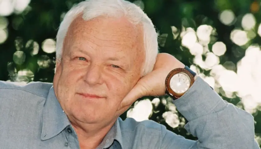 Uśmiechnięty Marian Opaniak, z siwymi włosami i niebieskimi oczami, opiera głowę na dłoni. Zegarek na jego nadgarstku wskazuje godzinę.