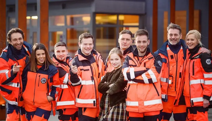 Uśmiechnięta na sygnale obsada w pomarańczowych kurtkach ratowniczych pozuje przed budynkiem.