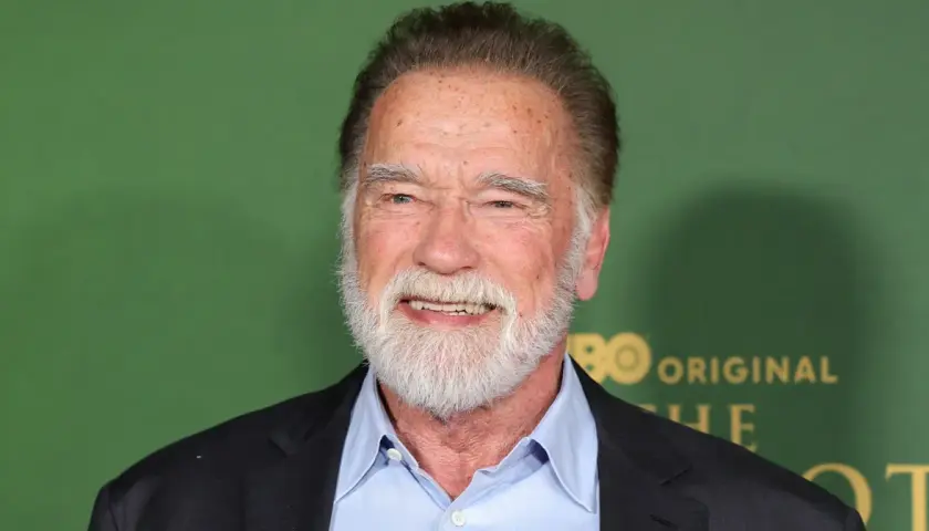 Arnold Schwarzenegger, z siwą brodą i w niebieskiej koszuli, uśmiecha się. W tle zielona ściana z logo HBO.