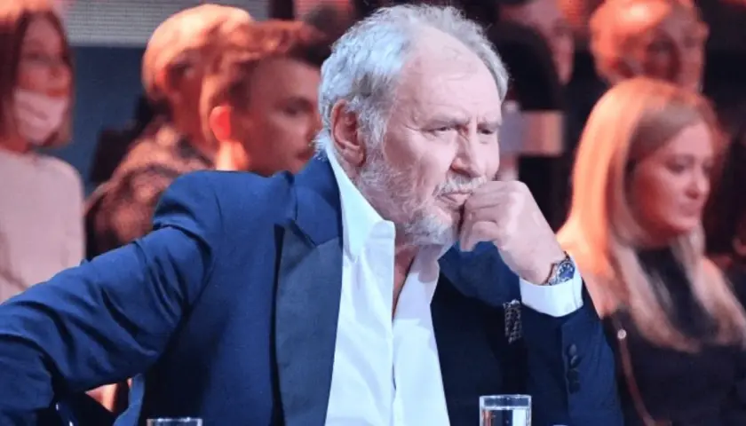 Andrzej Grabowski żyje, siedzi w eleganckim garniturze, zamyślony, z dłonią przy ustach. W tle widać innych ludzi.