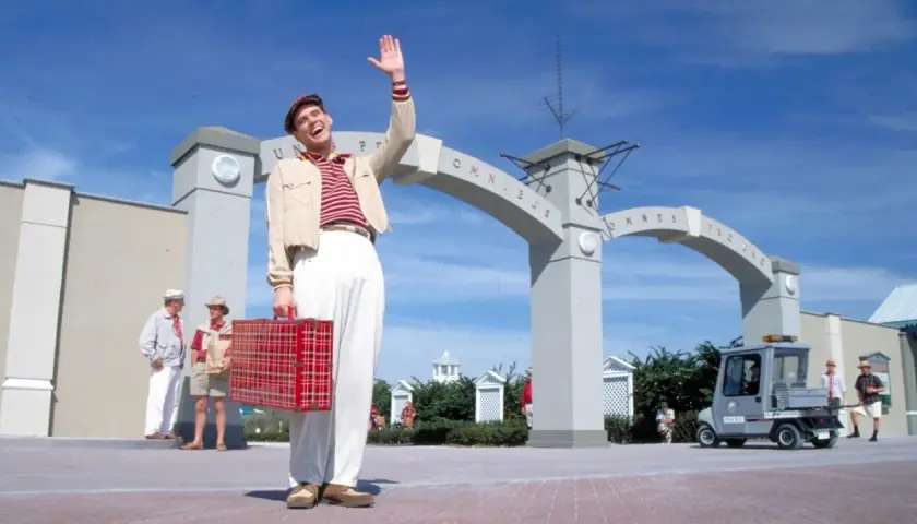 Truman Show obsada: Jim Carrey wita się z widzami, trzymając czerwoną walizkę. W tle łuk z napisem "Omnibus".
