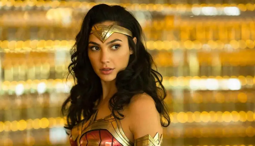 Gal Gadot jako Wonder Woman, część obsady filmu, w złotej zbroi i opasce na głowie, na tle rozmytych, złotych świateł.