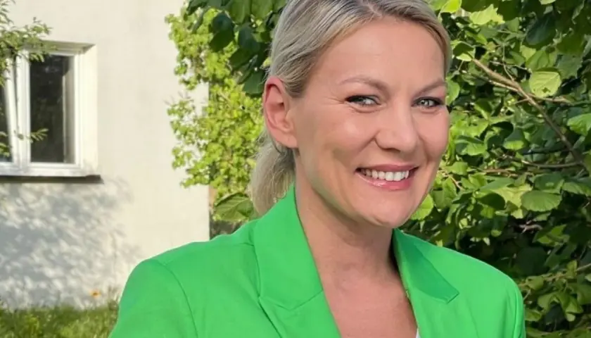 Uśmiechnięta Martyna Kupczyk w zielonej marynarce, na tle zieleni i białego domu.