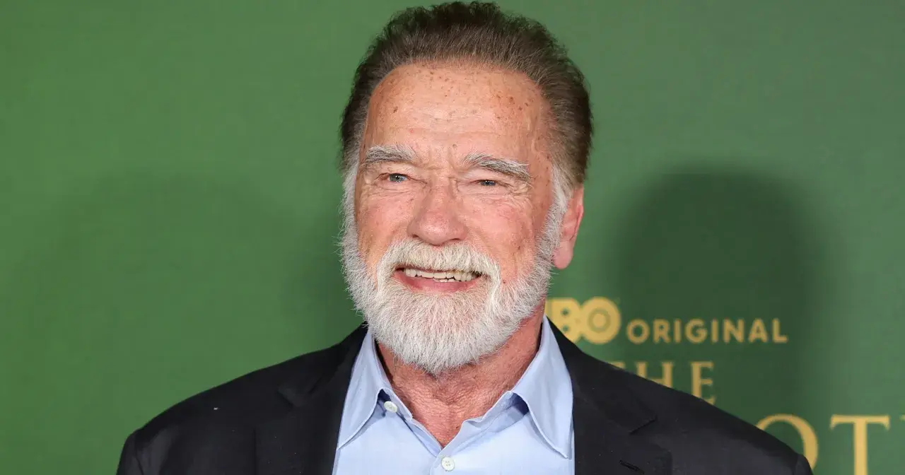 Arnold Schwarzenegger, z siwą brodą i w niebieskiej koszuli, uśmiecha się. W tle zielona ściana z logo HBO.