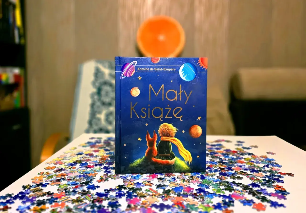 Odkryj, dlaczego warto przeczytać Małego Księcia. Książka z liskiem i chłopcem na tle kosmosu, otoczona rozsypanymi puzzlami.