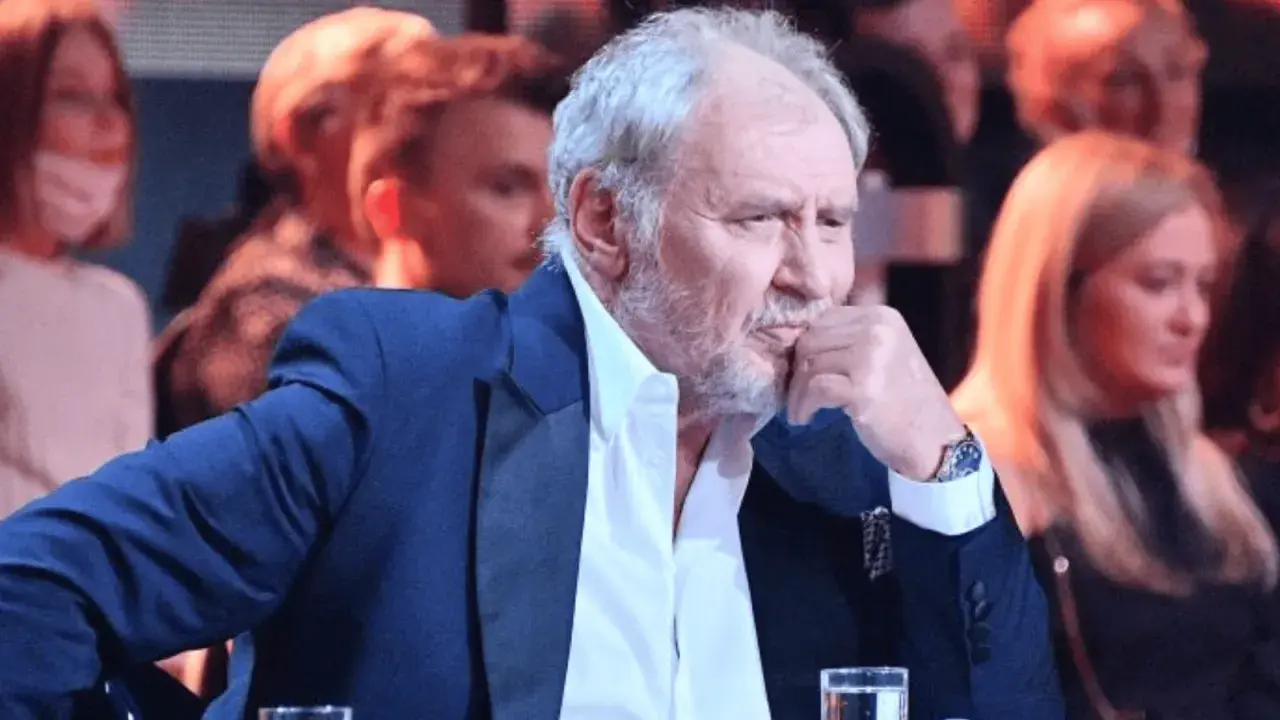 Andrzej Grabowski żyje, siedzi w eleganckim garniturze, zamyślony, z dłonią przy ustach. W tle widać innych ludzi.