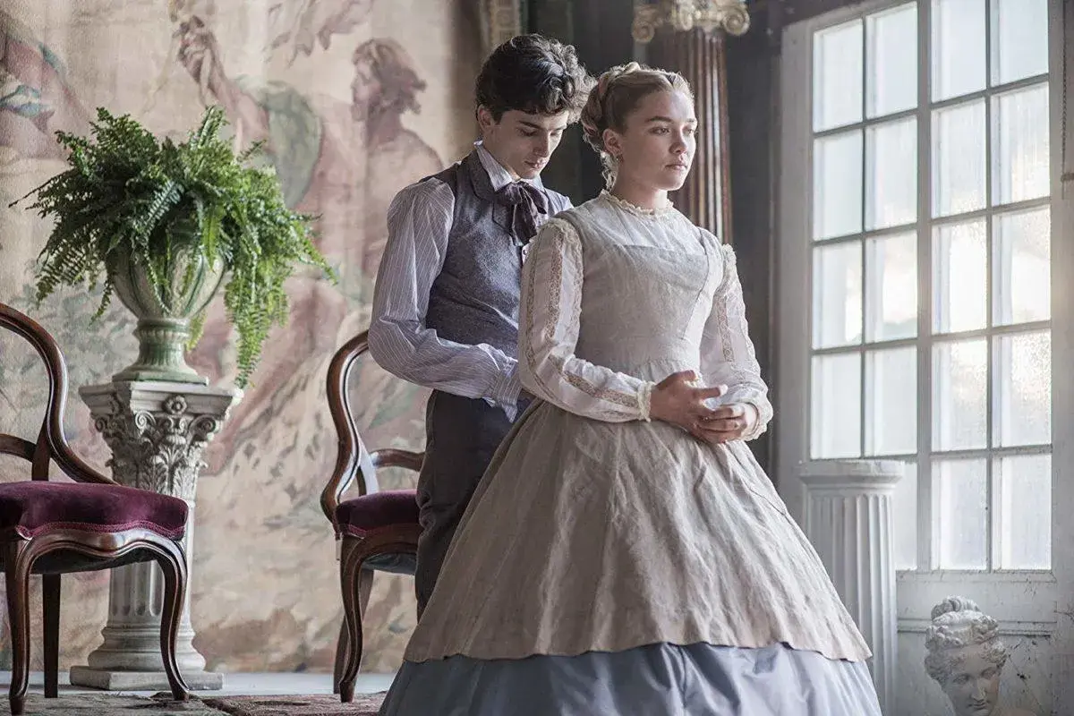 Małe kobietki obsada: Timothée Chalamet i Florence Pugh w strojach z epoki, w eleganckim wnętrzu.