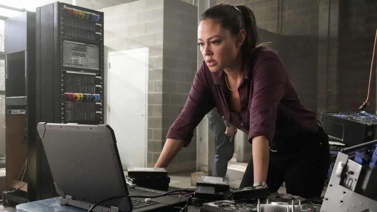 NCIS Hawaii obsada: Jane Tennant analizuje dane na laptopie, otoczona serwerami i dyskami twardymi.