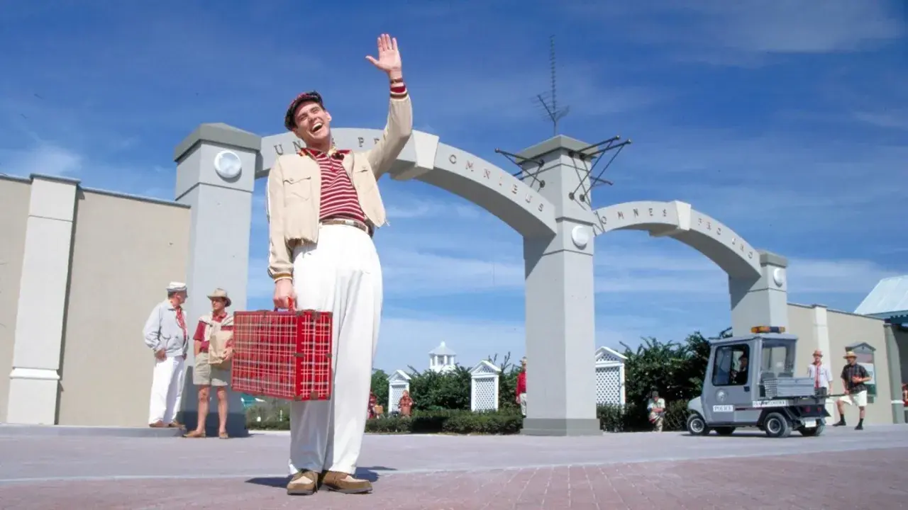 Truman Show obsada: Jim Carrey wita się z widzami, trzymając czerwoną walizkę. W tle łuk z napisem "Omnibus".