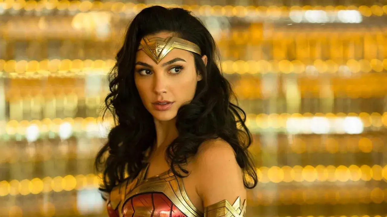 Gal Gadot jako Wonder Woman, część obsady filmu, w złotej zbroi i opasce na głowie, na tle rozmytych, złotych świateł.