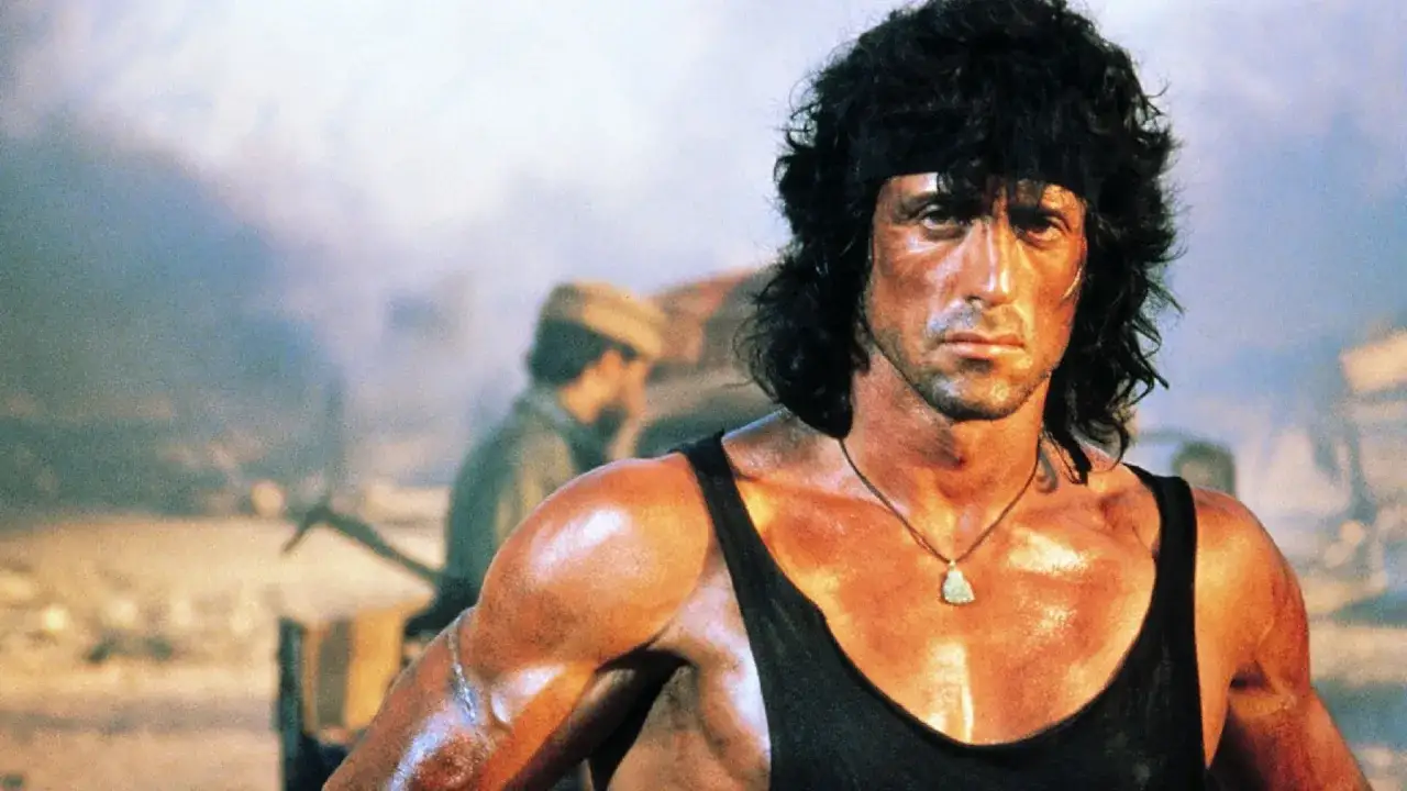 Sylvester Stallone jako Rambo, zdeterminowany, z opaską na czole. W tle dym i inni żołnierze. Ciekawe, ile lat ma Sylvester Stallone w tej roli?