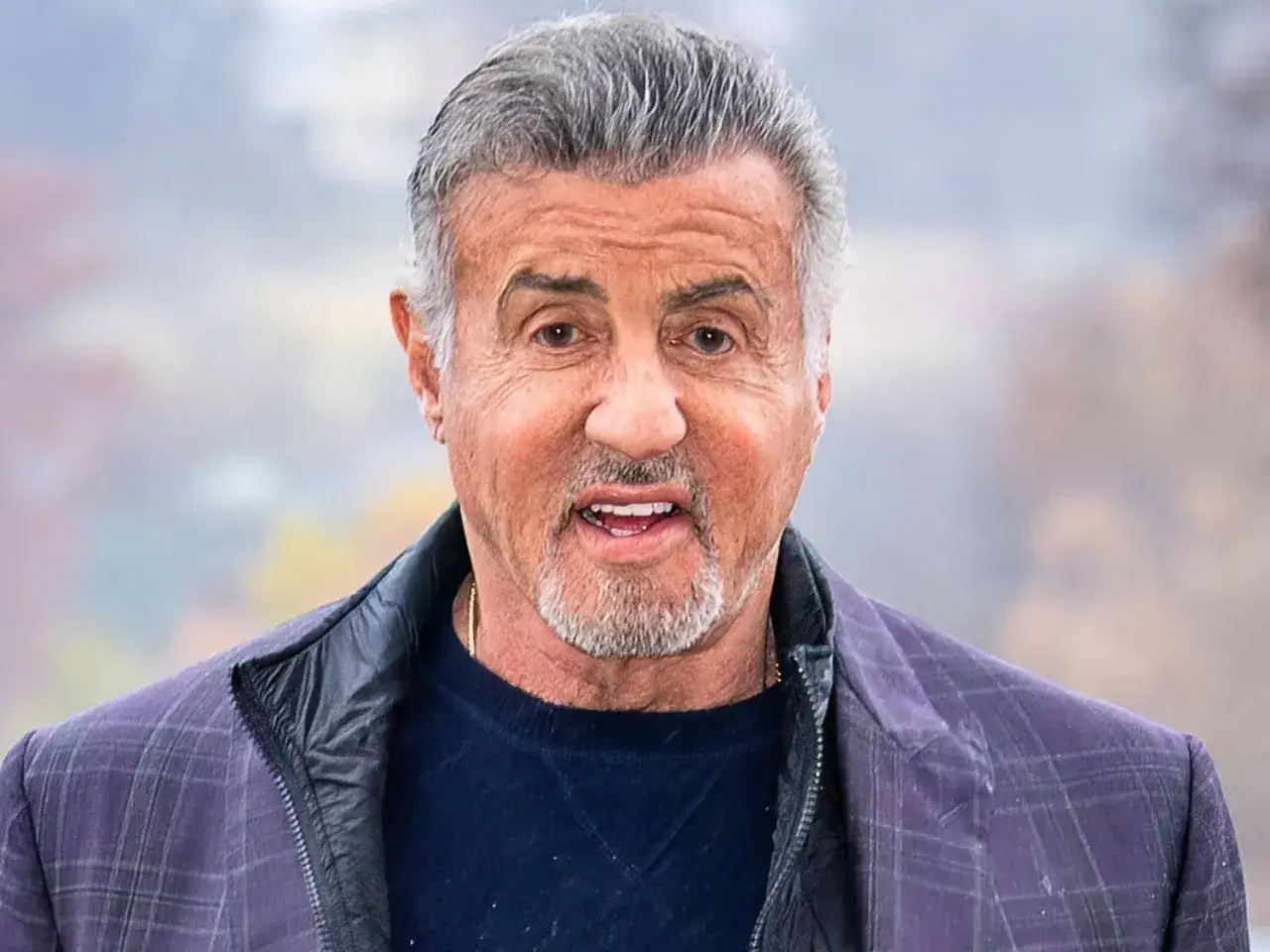Sylvester Stallone, z siwymi włosami i brodą, w kratowanej marynarce. Wiele osób zastanawia się, ile lat ma Sylvester Stallone.
