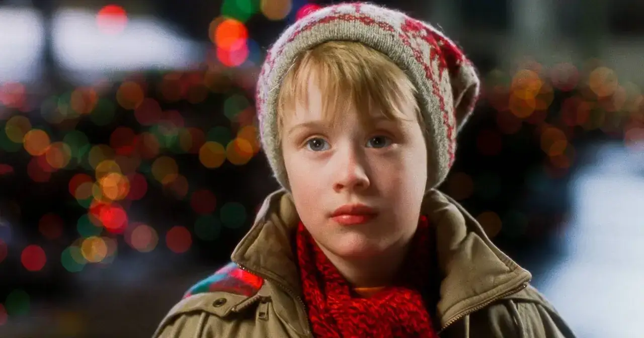 Macaulay Culkin, który grał Kevina samego w domu, w czapce i szaliku na tle świątecznych lampek.