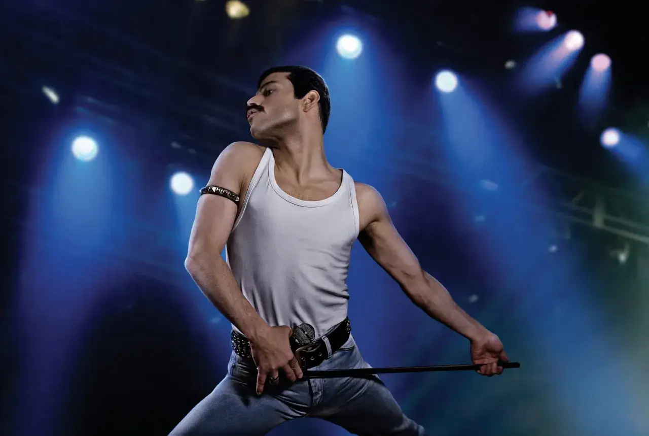 Rami Malek jako Freddie Mercury, w białej podkoszulce i dżinsach, z mikrofonem przy pasie, śpiewa w