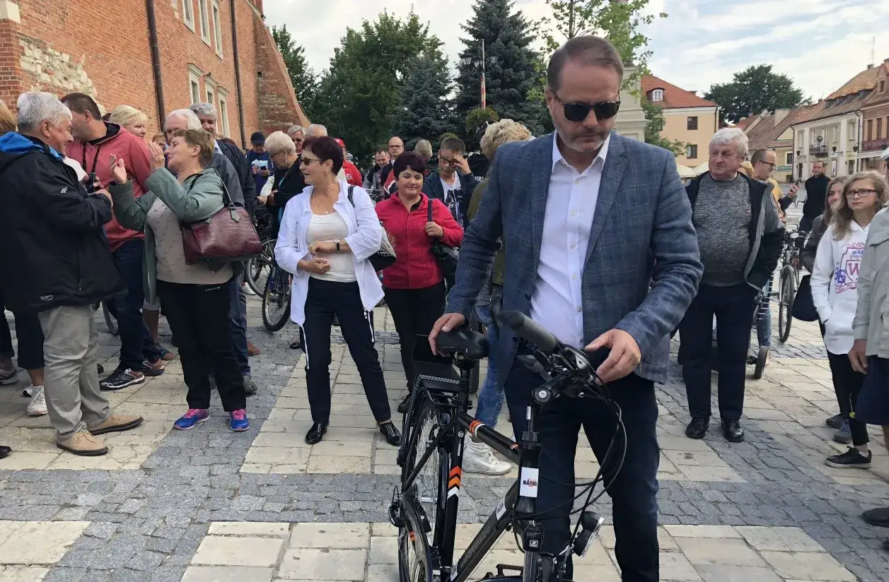 Mężczyzna w okularach, który wygląda jakby grał Ojca Mateusza, stoi obok roweru wśród tłumu ludzi na brukowanym placu.