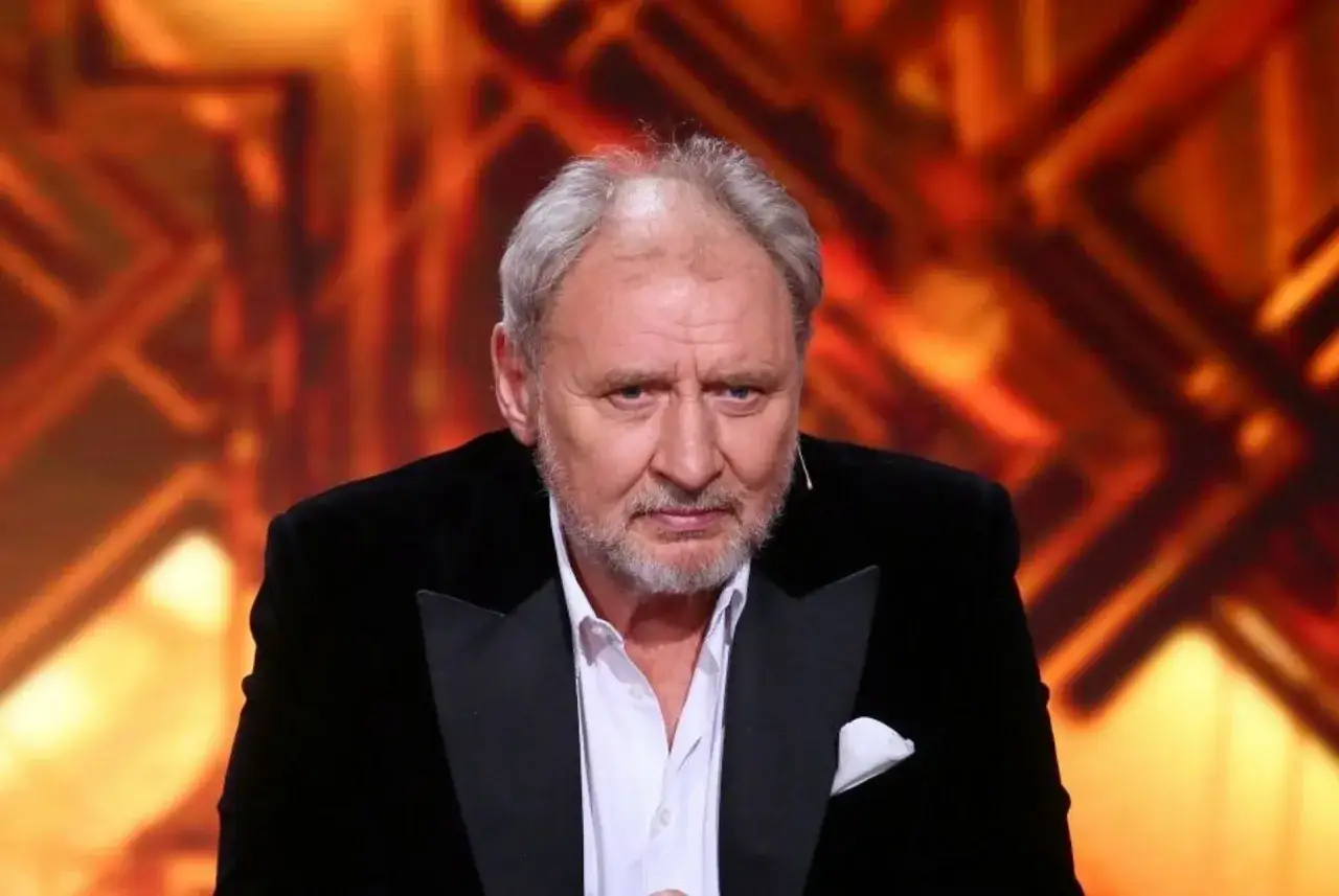 Andrzej Grabowski żyje i wygląda na zmęczonego, siedząc w czarnym smokingu na tle pomarańczowej scenografii.