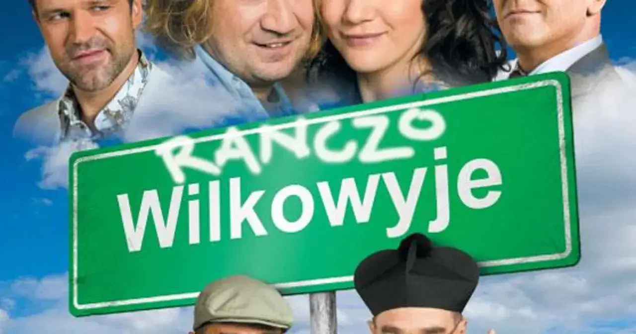 Uśmiechnięta obsada serialu Ranczo Wilkowyje na tle nieba i chmur, z charakterystycznym zielonym znakiem drogowym.