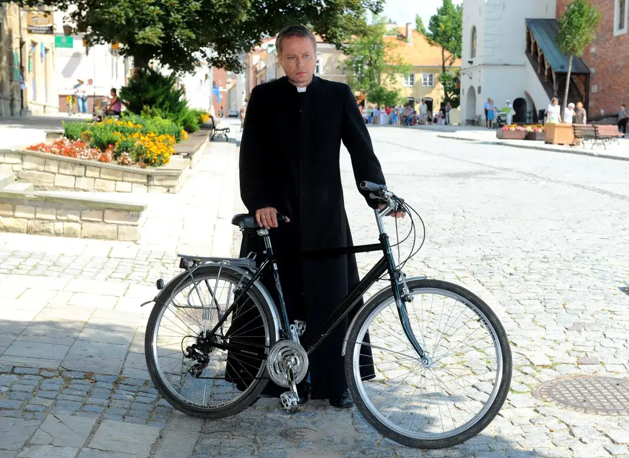 Aktor, który gra Ojca Mateusza, w czarnej sutannie stoi obok roweru na brukowanej ulicy.
